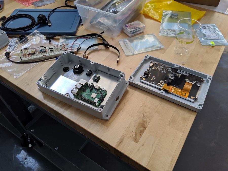 A device enclosure and lid sit on a workbench, with PCB assemblies mounted inside. Various connectors and cables are also on the benchtop.