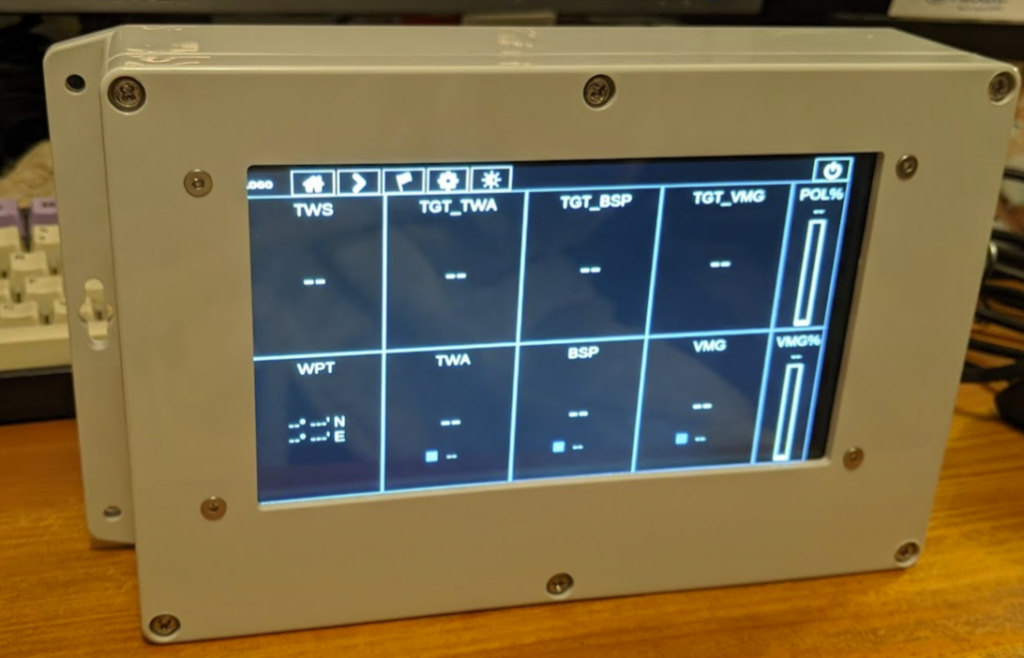 An embedded hardware device of a computer with an LCD inside a plastic enclosure sits on a desk. A dashboard of various (blank) instrument readings and calculations is shown on the LCD.