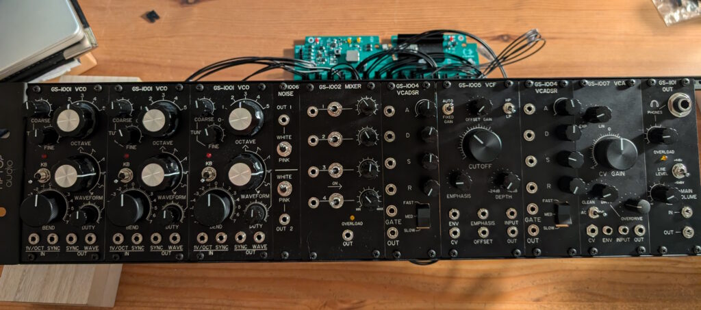 A collection of Eurorack modules arranged side-by-side on rack rails so that you can see their front panels with knobs, switches, and jacks.

The modules, from left to right: 3 VCO's, a noise generator, a 4-channel mixer, VCADSR, VCF, VCADSR, VCA, and a headphone amplifier / line out module.

The rack of modules is sitting on a table top; another PCB is visible on the table as well, with wires connecting it to the rear side of the Eurorack modules.