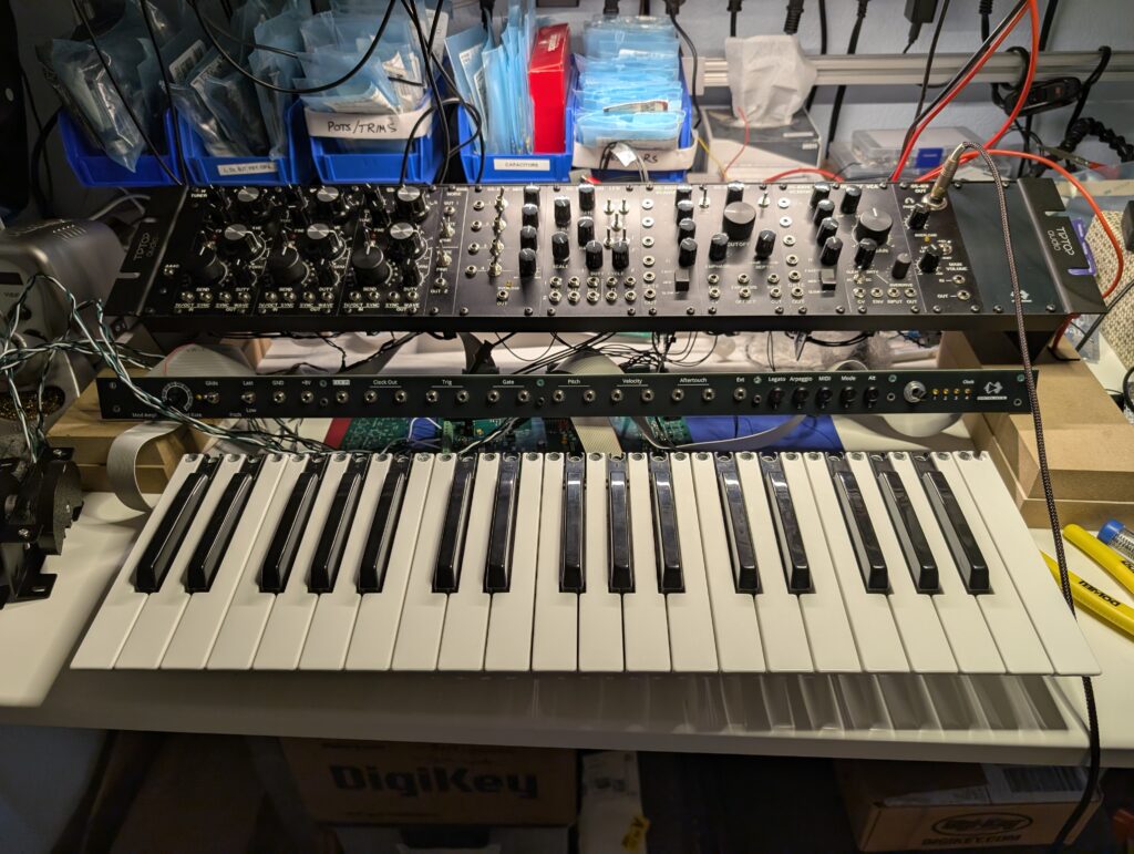 A collection of Eurorack modules, PCBs, components with front panels, and a prominent 37-key piano keyboard spread out on a workbench and wired together.